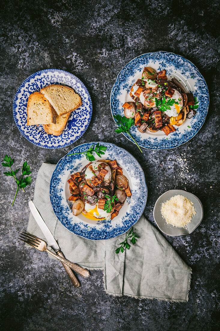 Two pretty antique plates with runny poached eggs, shallots, mushrooms and bacon in red wine sauce, with a plate of toast, on a dark background