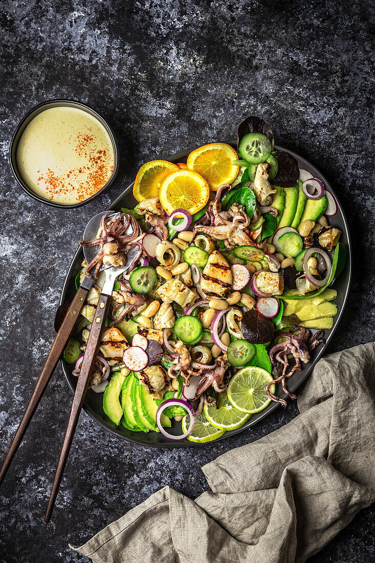 Grilled squid salad with orange, lime and avocado on a dark platter with sauce bowl