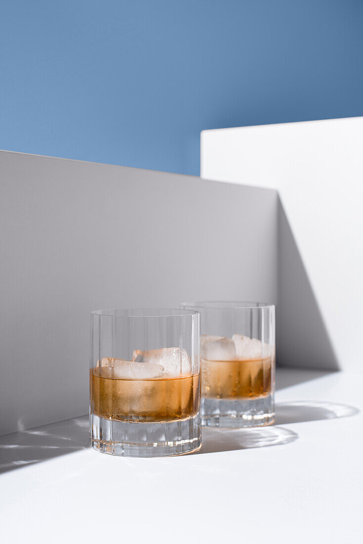 Close up of translucent glasses filled with cold refreshing scotch whiskey with ice cubes placed on white surface against white wall