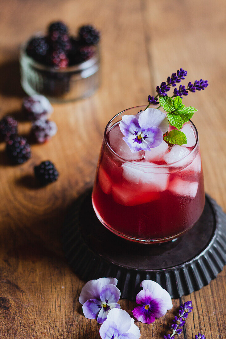 Bramble cocktail with violet flower, mint and lavender on a wood backdrop