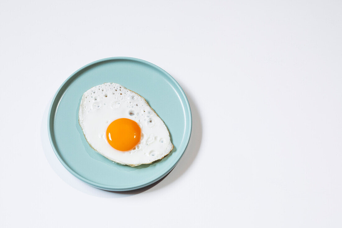 Top view of a delicious breakfast with fried egg with yellow egg yolk, served on a blue ceramic plate on a table against a white background