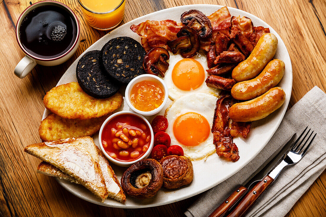 Ein komplettes englisches Frühstück mit Spiegeleiern, Würstchen, Speck, Blutwurst, Bohnen, Toast und Tee auf Holzuntergrund