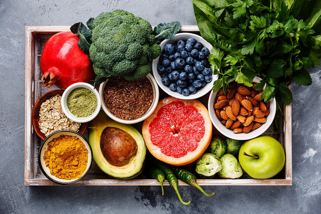 Gesunde Lebensmittel Clean Eating Auswahl in Holzkiste: Obst, Gemüse, Samen, Superfood, Getreide, Blattgemüse auf grauem Beton Hintergrund