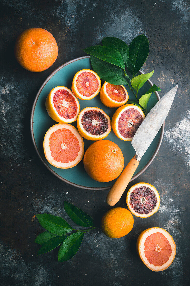 Blutorangen und rosa Grapefruit, halbiert und ganz, mit Zitrusblättern, in blauer Keramikschale und auf Tisch, mit Messer auf dunklem Hintergrund