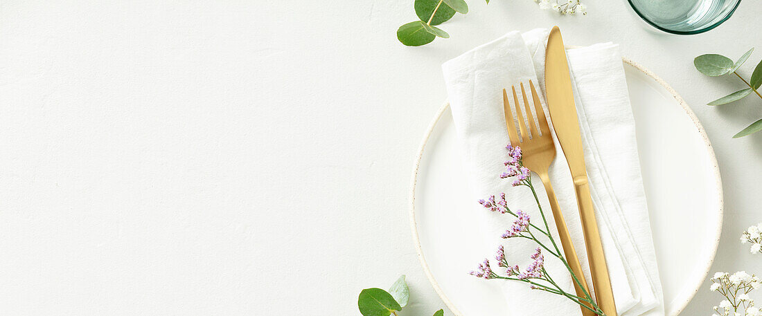 Gold cutlery with eucalyptus branches on a white plate with a napkin against a light grey background. Minimalist design. Space for copying