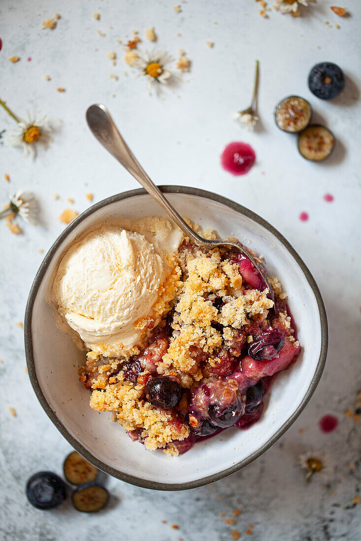 Apple and blackberry crumble in a bowl with a scoop of vanilla ice cream. A classic spoon is wedged into the bowl