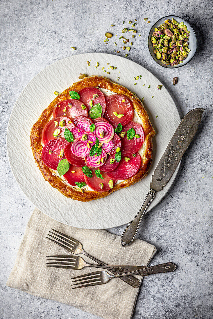 Round puff pastry cake with sliced beetroot, feta and mint leaves on a white plate with pistachios in shells