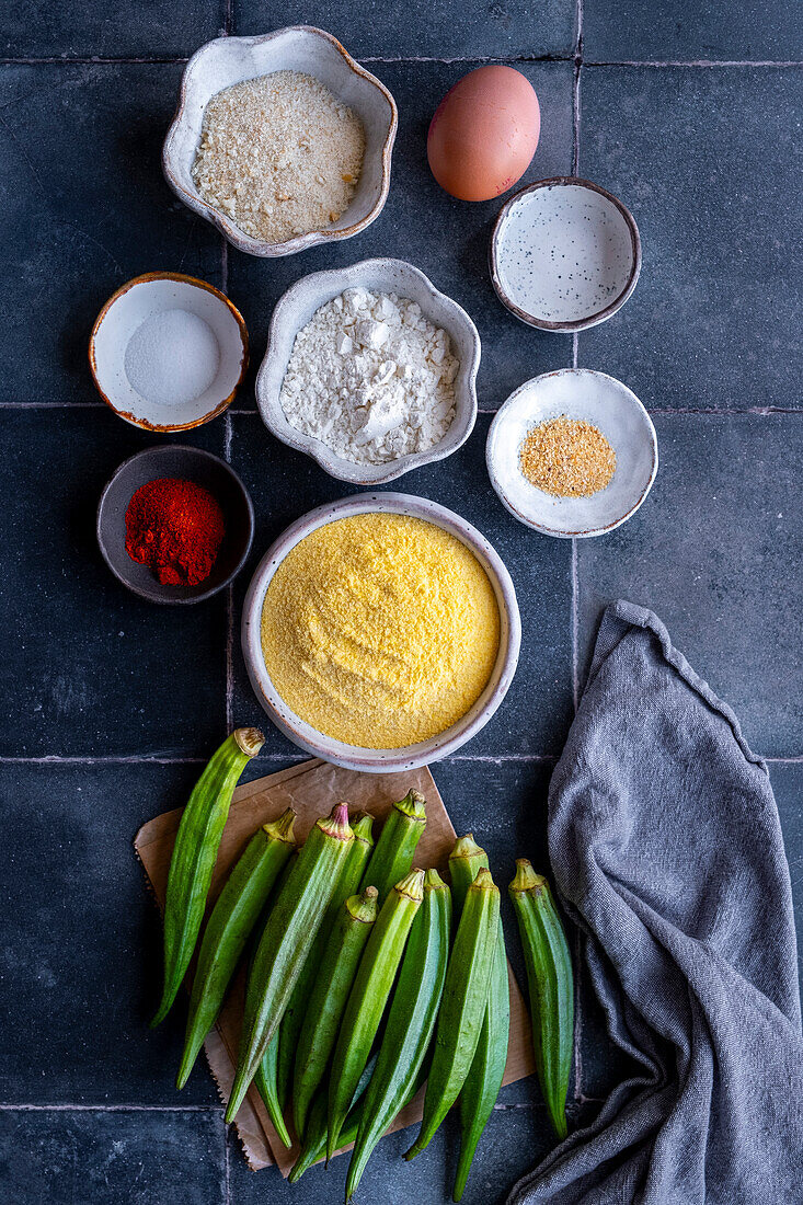 Fresh okra, cornmeal, flour, bredcrumbs, egg, water, salt, paprika and garlic powder on a grey backdrop.