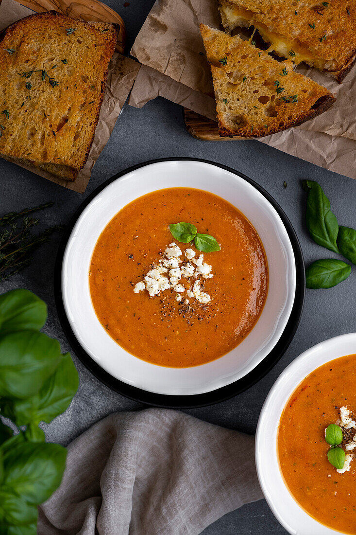 Roasted tomato soup topped with feta cheese and basil leaves in two white bowls and grilled cheese sandwiches on the side.