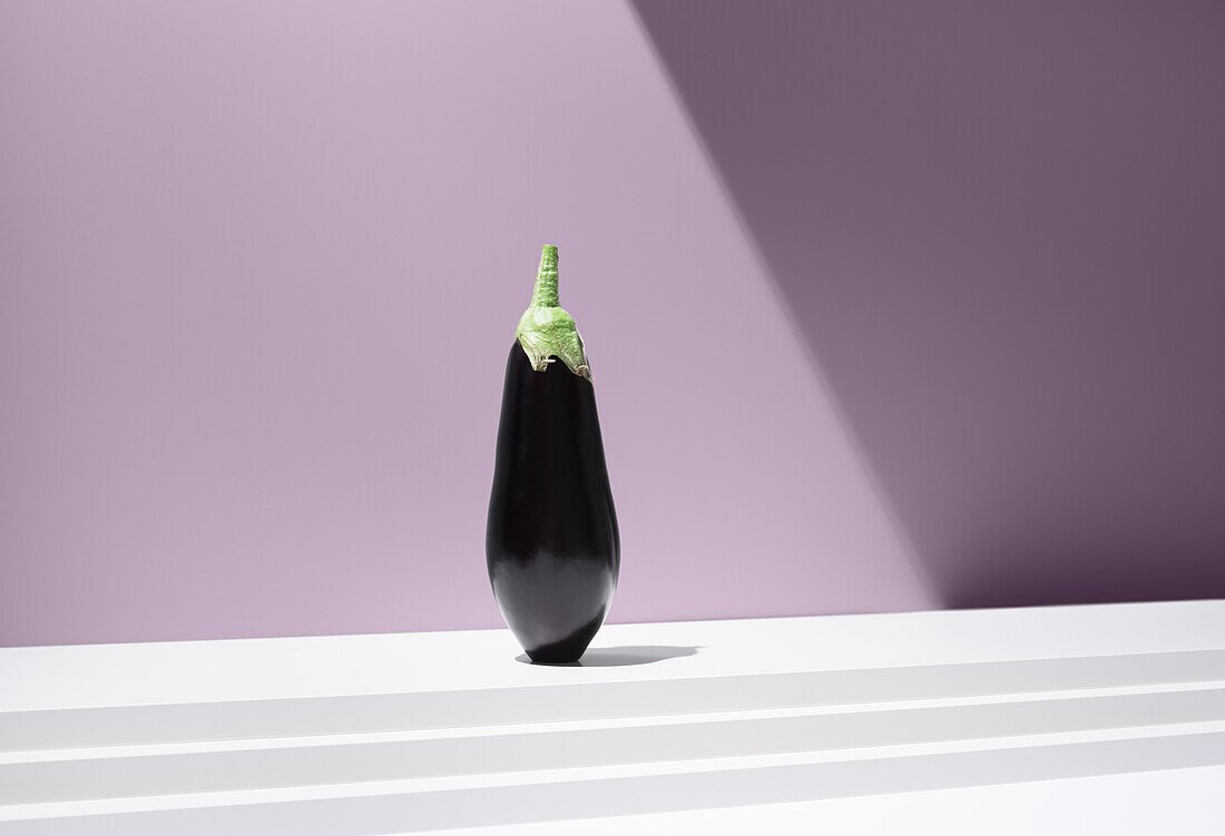 Vivid image of a black aubergine with a green cap on a white, stepped plate against a purple background under bright light