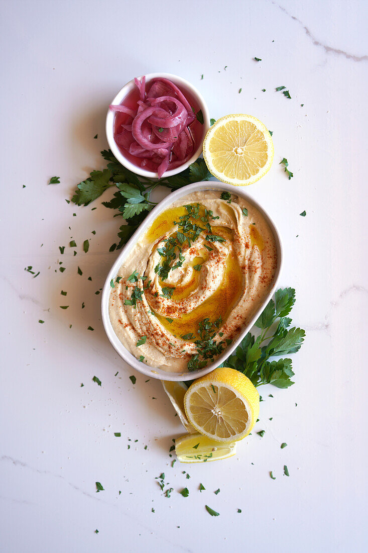 Kichererbsen-Hummus, serviert mit Olivenöl, eingelegten roten Zwiebeln, Zitronen und Fladenbrot auf einem weißen Marmortisch. Weibliche Hand dippt Fladenbrot