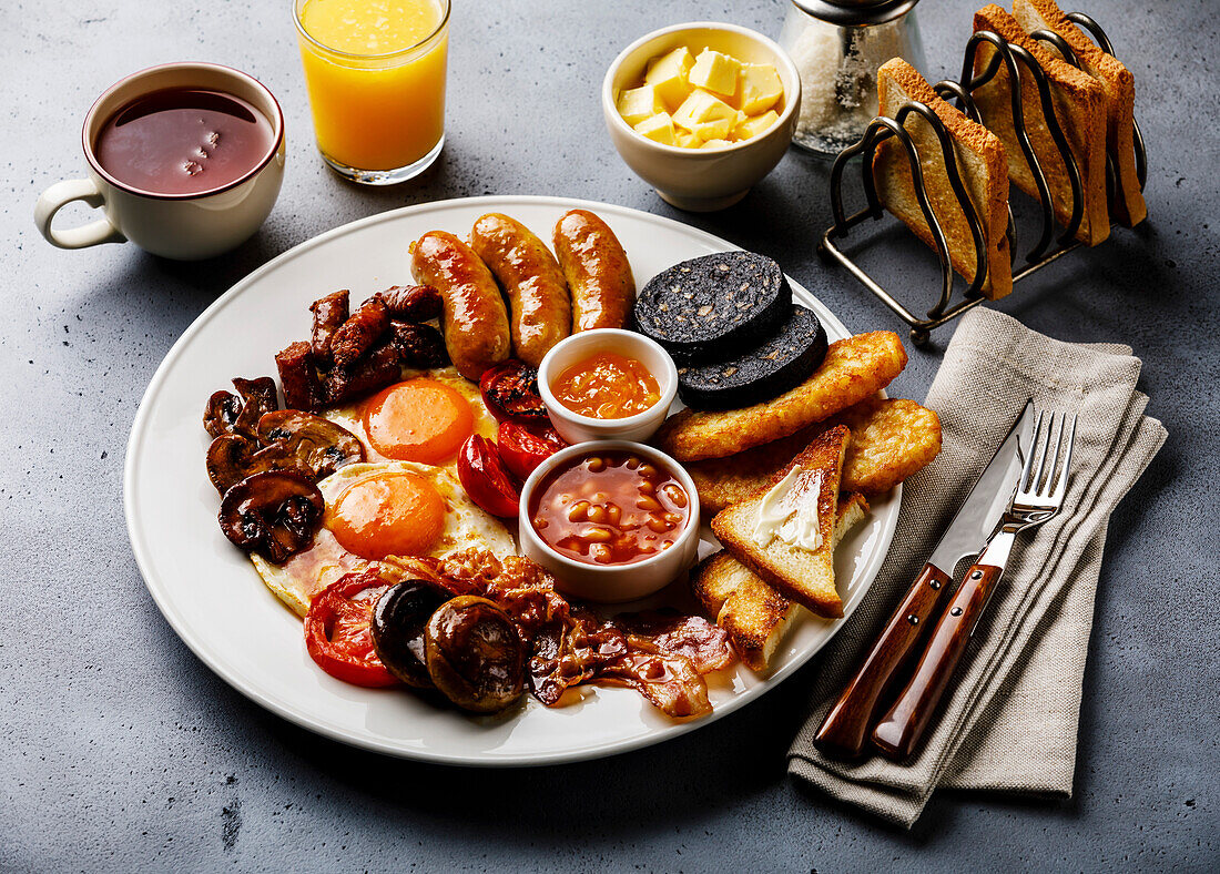 Ein komplettes englisches Frühstück mit Spiegeleiern, Würstchen, Speck, Blutwurst, Bohnen, Toasts und Tee auf grauem Betonhintergrund