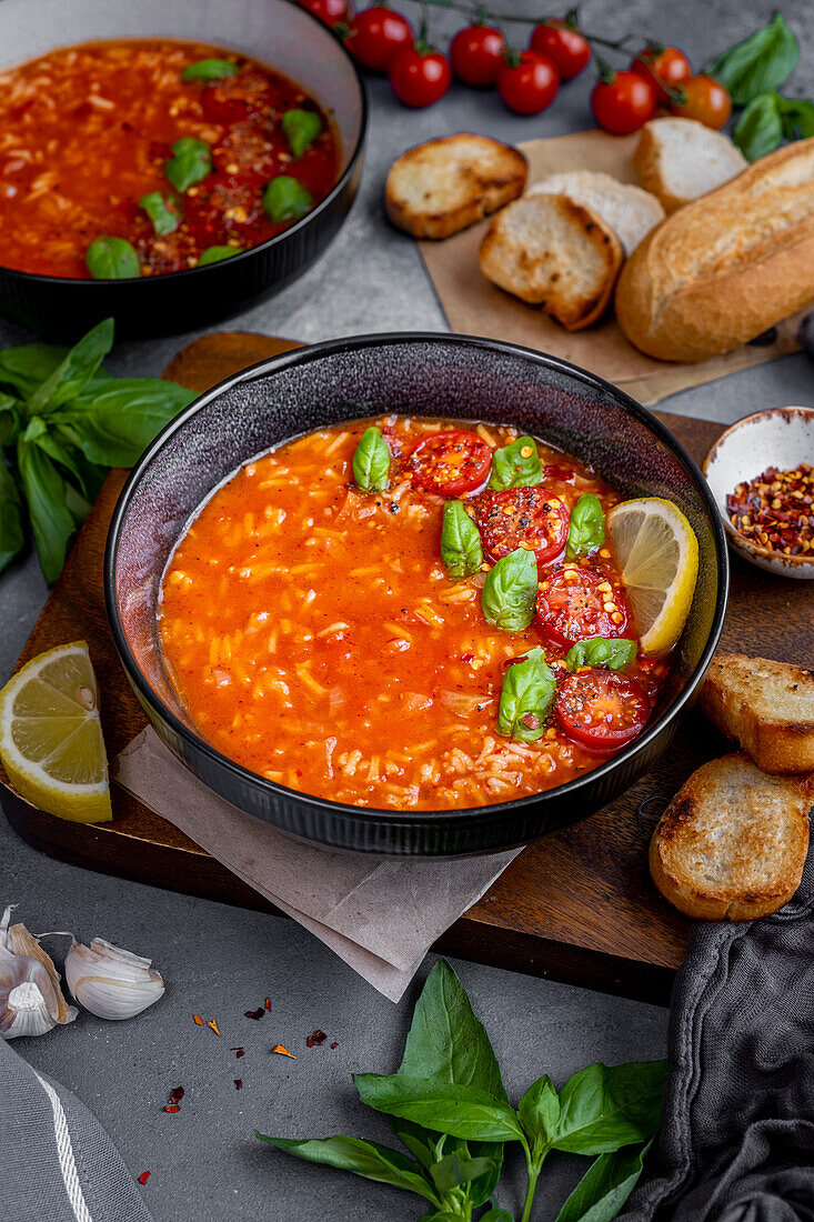 Tomaten-Reis-Suppe garniert mit Basilikumblättern, Kirschtomaten und einer Zitronenscheibe in einer schwarzen Schale, dahinter Brotscheiben, Basilikumblätter und eine weitere Schale mit Suppe