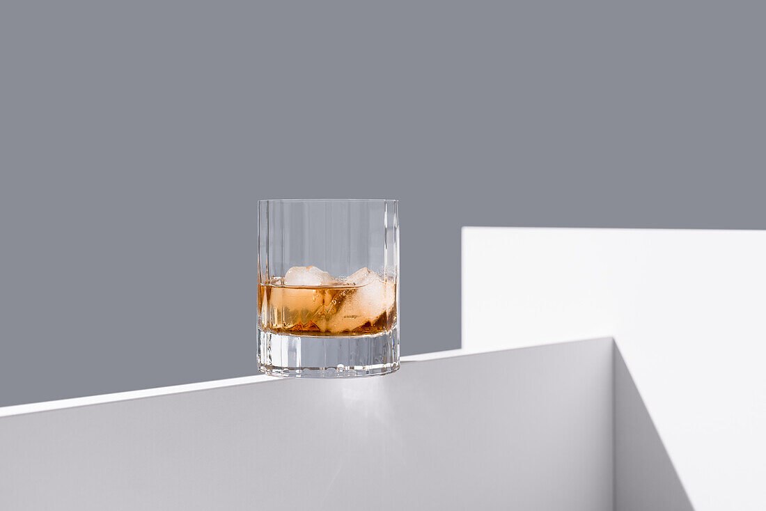 Transparent glass of whiskey on rocks with ice cubes on white wooden table isolated on grey background