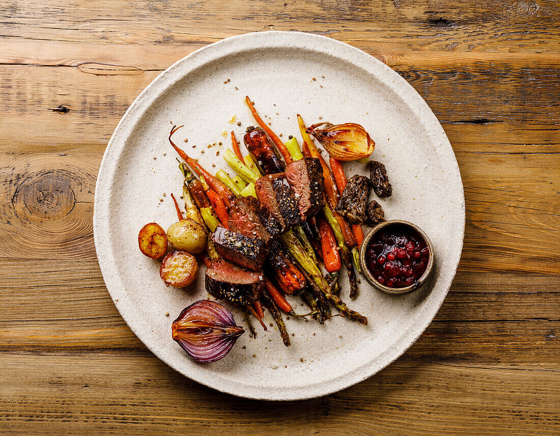 Grilled sliced Venison Steak with baked vegetables and berry sauce on wooden background