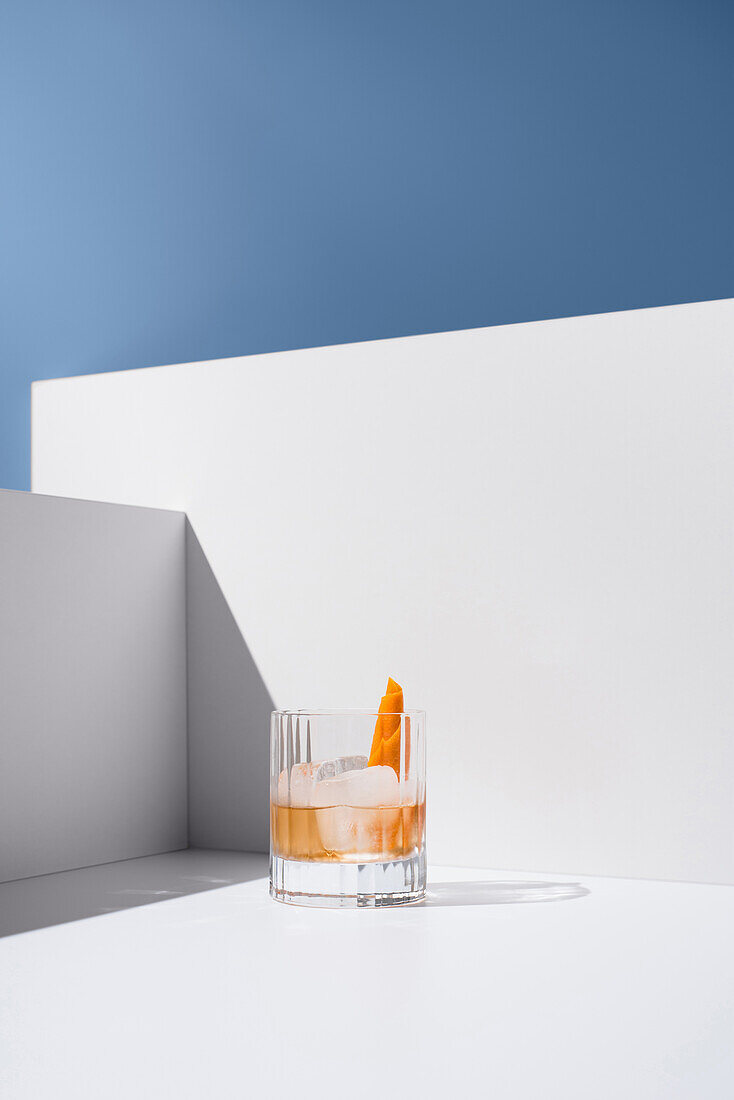 Crystal clear glass filled with fresh old-fashioned cocktail garnished with oranges and ice cubes on a white surface between white walls against a grey background