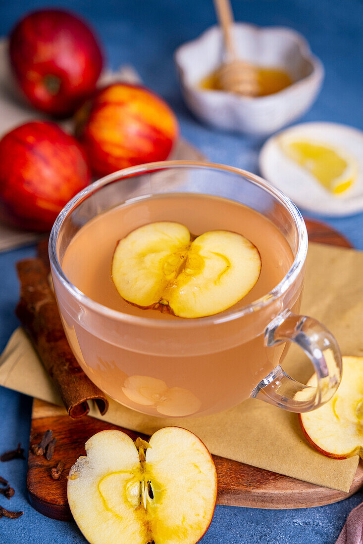 A cup of homemade apple tea topped with a slice of apple and more red apples, honey and a lemon slice behind it.
