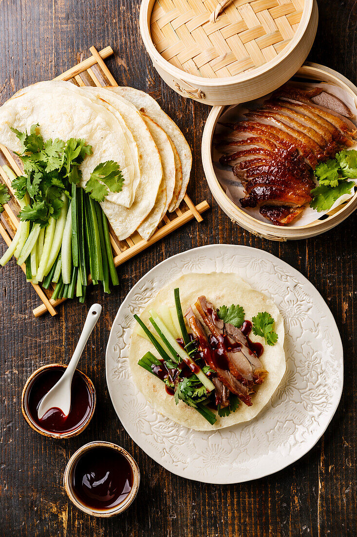 Peking duck in portion size with fresh cucumber, green onion, coriander and toasted Chinese wheat pancakes with Hoysin sauce on a wooden base