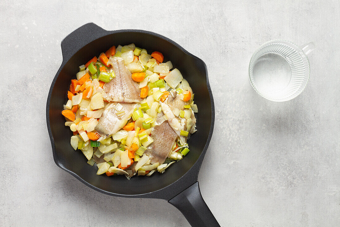 Top view of a frying pan with fumet containing raw sliced fish and various sliced carrot and cabbage vegetables on a grey table background