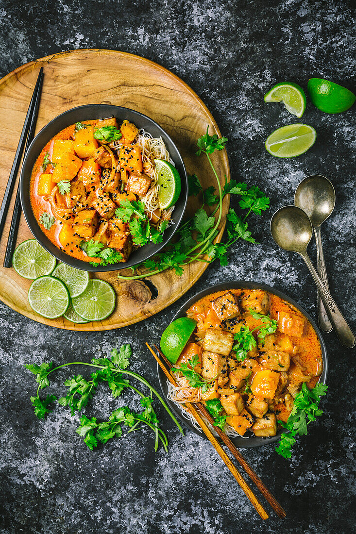 Two bowls of red coconut curry with tofu and butternut squash with rice noodles, in dark bowls on a wooden board, with lime slices and fresh coriander
