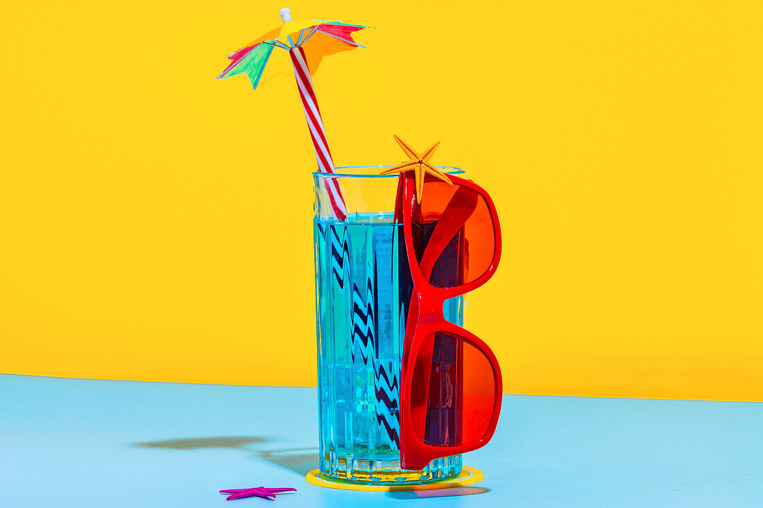 Front view of a composition of a glass of blue liquid with a straw and stylish red glasses on a light blue and yellow background in the studio