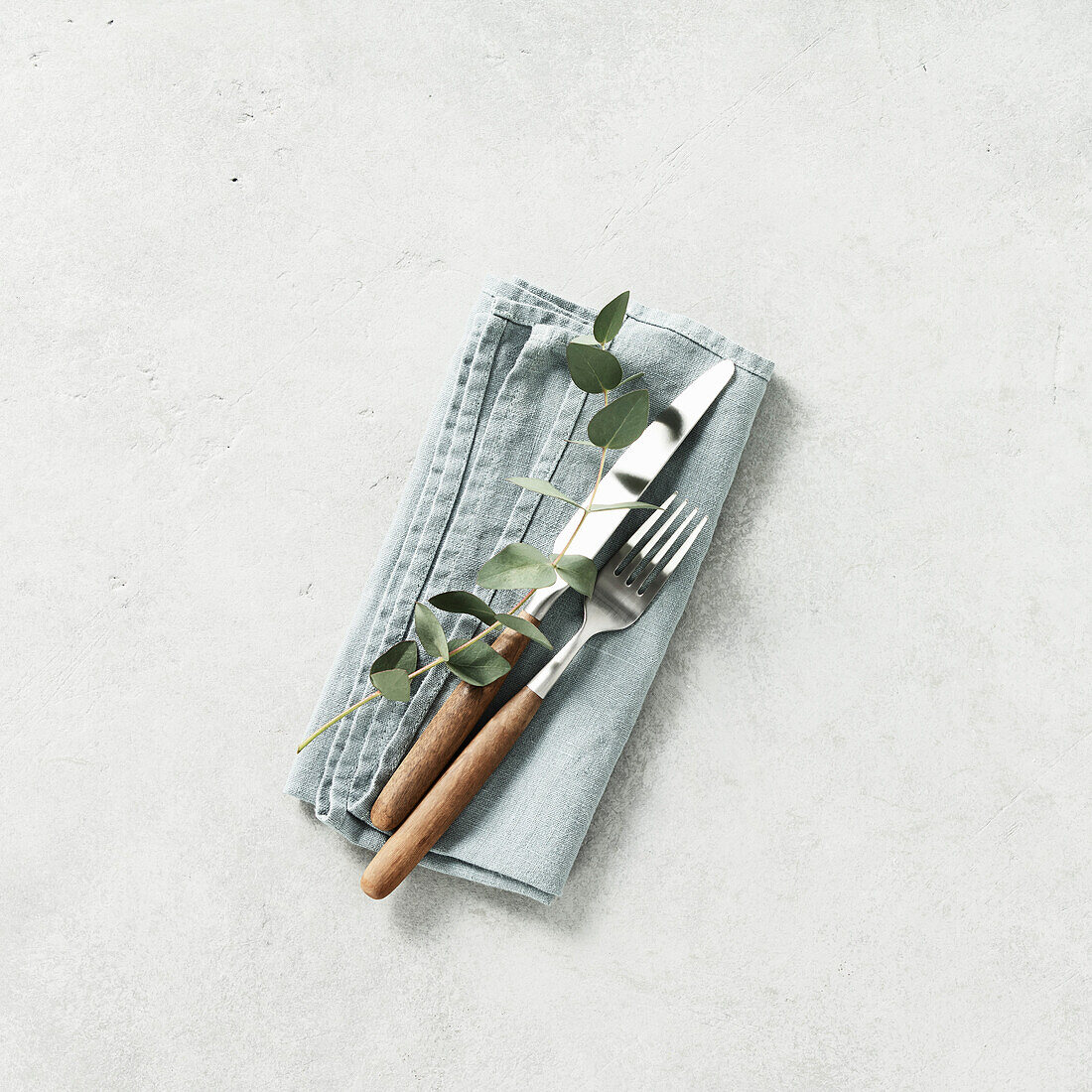 Gedeckter Tisch mit Leinenserviette, Gabel, Messer und Eukalyptus auf grauem Hintergrund Quadratische Komposition