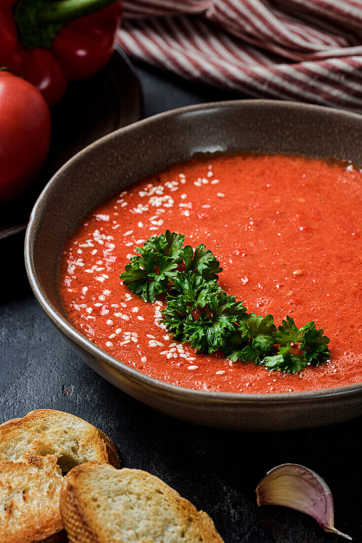 Gazpacho garniert mit Petersilie in einem Teller mit gewelltem Rand. Auf dem Tisch liegen rote Paprika, Tomaten, Knoblauch und Croutons. Ansicht von oben