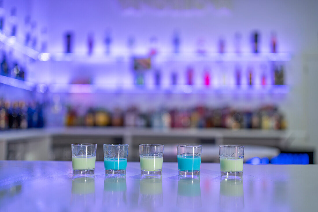 Row of served shots with blue and white spirits placed in row on counter against blurred shelves with bottles in restaurant bar