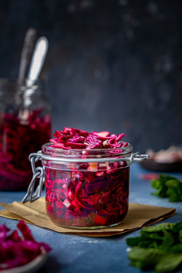 A small glass with pickled red cabbage and a larger one behind it, surrounded by garlic cloves and herbs
