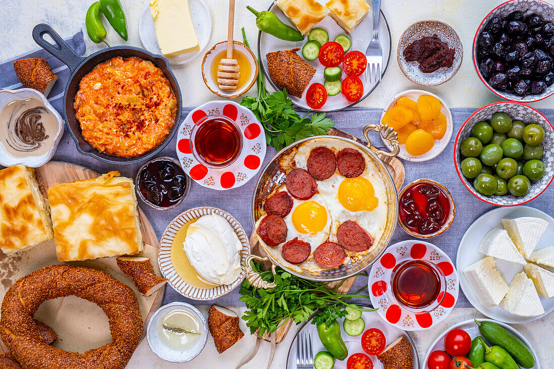 Flat-lay of Turkish breakfast with egg dishes in copper pan, pastries like borek and simit, jams, olives, cheese, vegetables and Turkish tea in traditional tulip glasses.