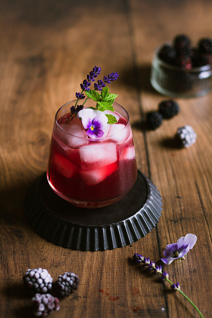 Brombeer-Cocktail mit Veilchenblüten, Minze und Lavendel vor einem Holzhintergrund