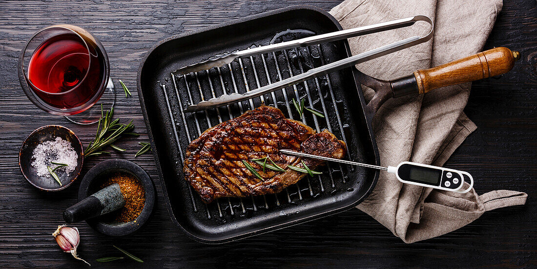 Gegrilltes Steak Striploin auf Pfanne und Fleischthermometer auf schwarzem verbranntem Holzhintergrund
