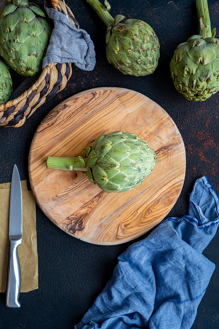 An artichoke on a round wooden cutting board, a knife and more artichokes accompany.