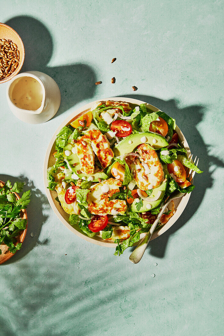 Halloumi and avocado tomato salad with sunflower seeds, yoghurt dressing, herbs and water on a green background with shade