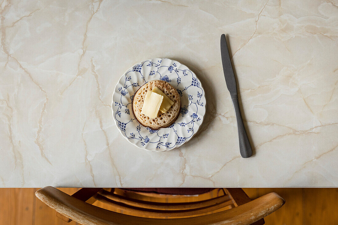 Crumpet with butter on blue floral plate on a marble table.