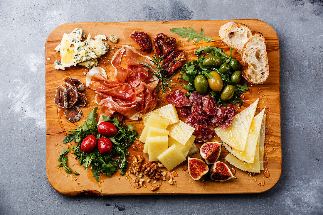 Italian snacks with ham, olives, cheese, sun-dried tomatoes, sausage and bread on a wooden chopping board against a concrete background