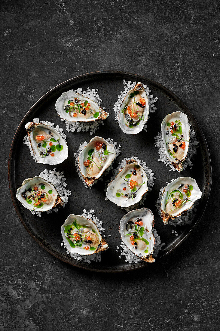 Frisch geschälte Austern mit Buttermilchdressing, Lachsrogen, Balsamico-Perlen und Kräuteröl auf Steinsalz auf einem schwarzen Teller