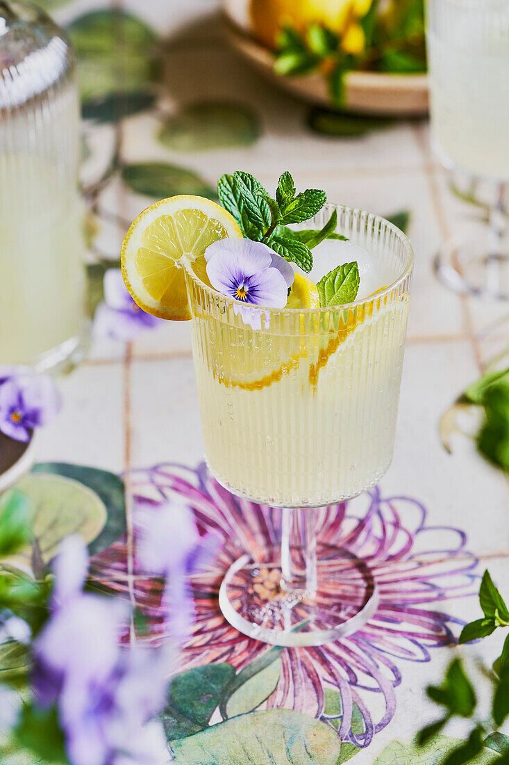 Zitronen-Minze-Limonade Mocktail auf floralem Hintergrund mit Zitrone, Minze und lila Blumendekor