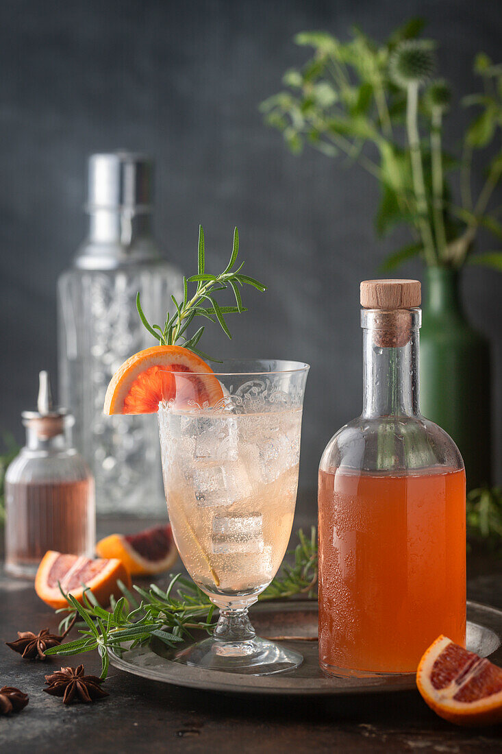 Light-colored cocktail on ice with blood orange syrup and club soda on ice in vintage glass with syrup bottle, citrus and rosemary garnish