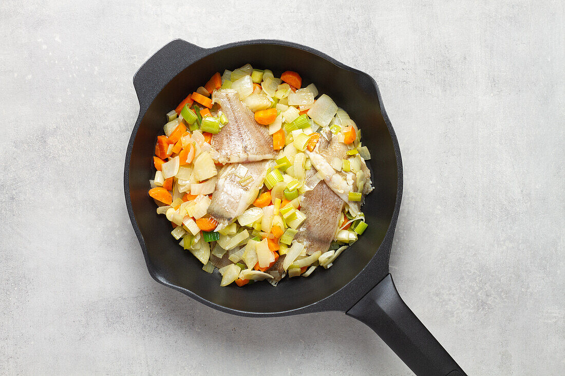 Top view of frying pan with fumet containing raw sliced fish and various sliced carrot and cabbage vegetables on grey table background