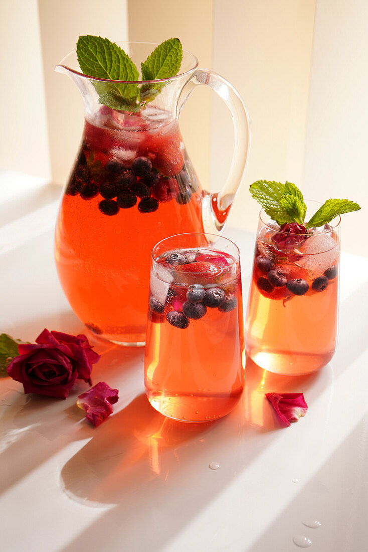 Beeren- und Rosenblütenschorle erfrischendes Sommergetränk auf weißem Tisch. Nahaufnahme von Krug und zwei Gläsern