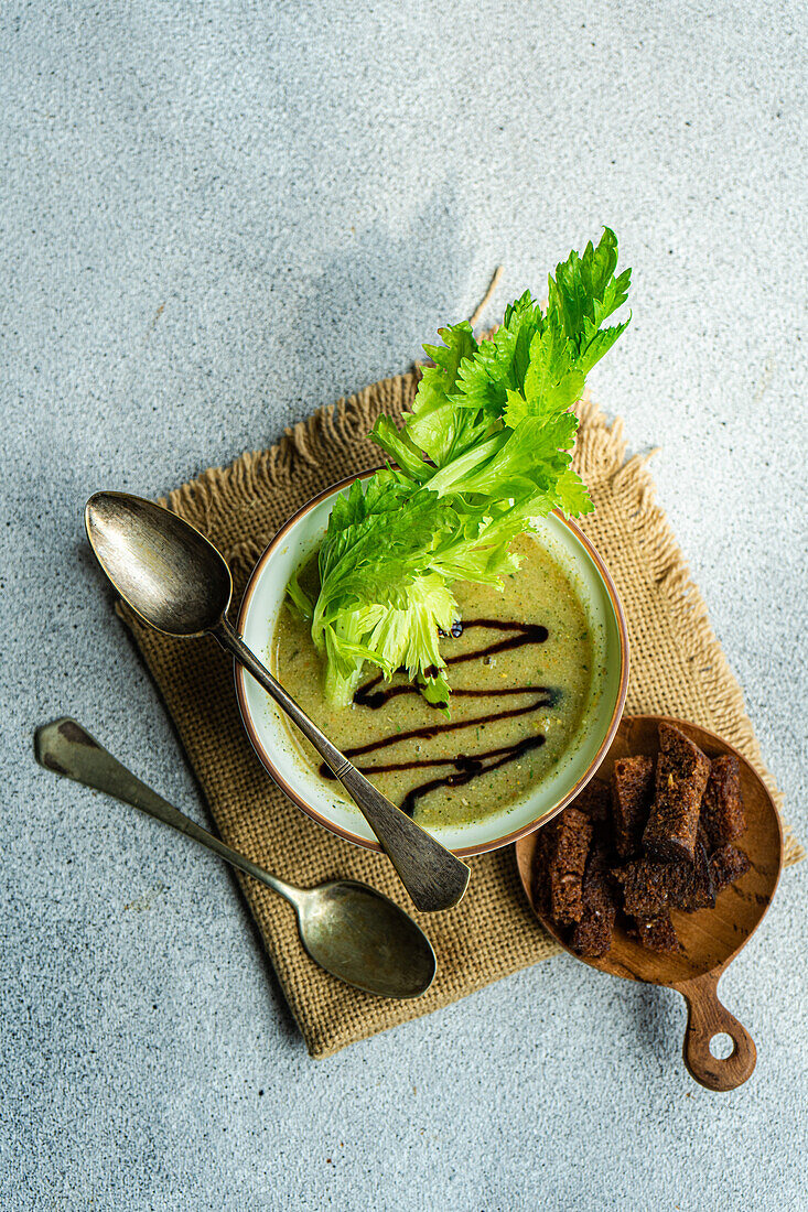 Top view of healthy celery cream soup in bowl with celery sticks and bowl with slices of bread placed on napkin with spoons against grey background