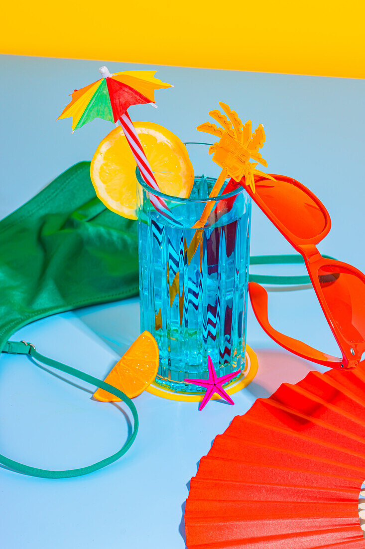 Composition of glass of cocktail and sliced orange fruit with straw, red glasses over surface with inner wear, hand fan on blue and yellow background