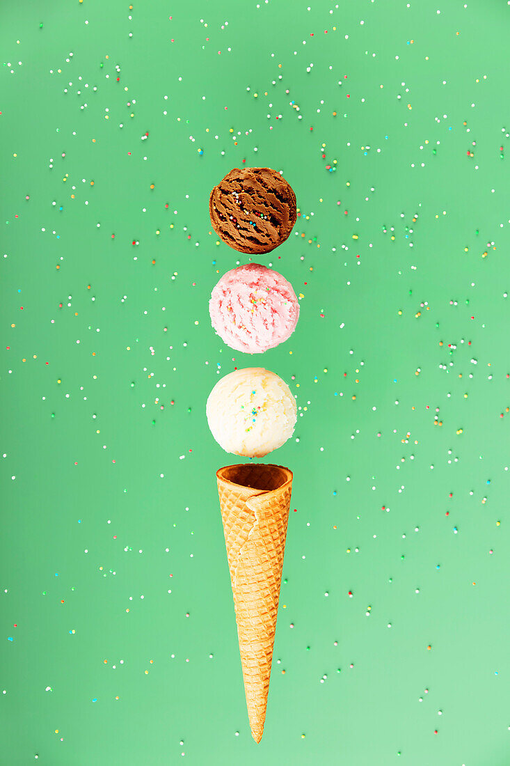 Chocolate, vanilla, strawberry balls and waffle cone on a green background with colourful sprinkles, floating concept. Spring or summer mood