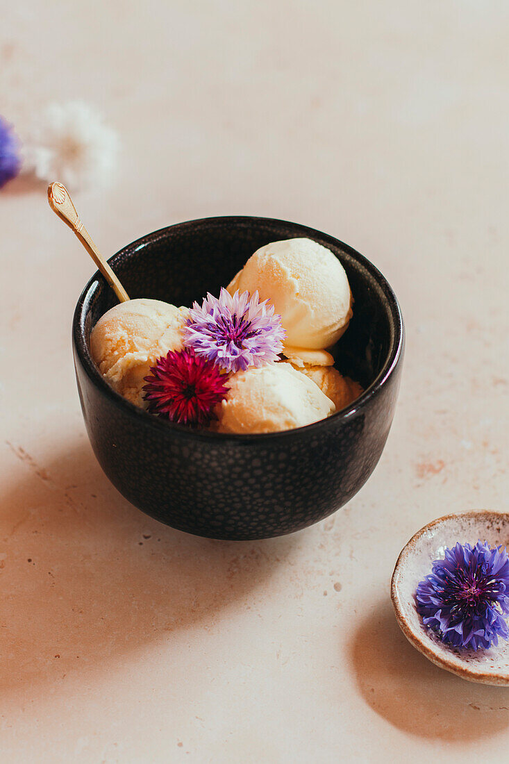 Vanilla ice cream scoops with edible flowers in a black bowl on a light-coloured background