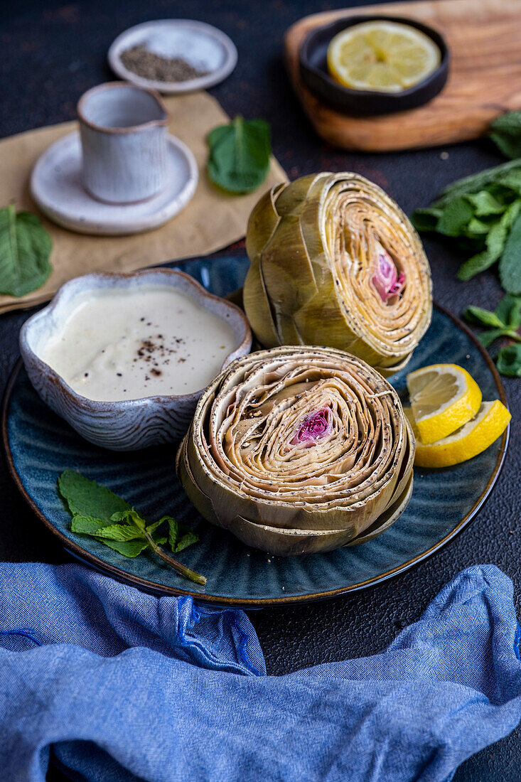 Steamed artichokes garnished with lemon slices and fresh mint leaves served with a bowl of a dipping sauce on a dark blue plate photographed from front view.