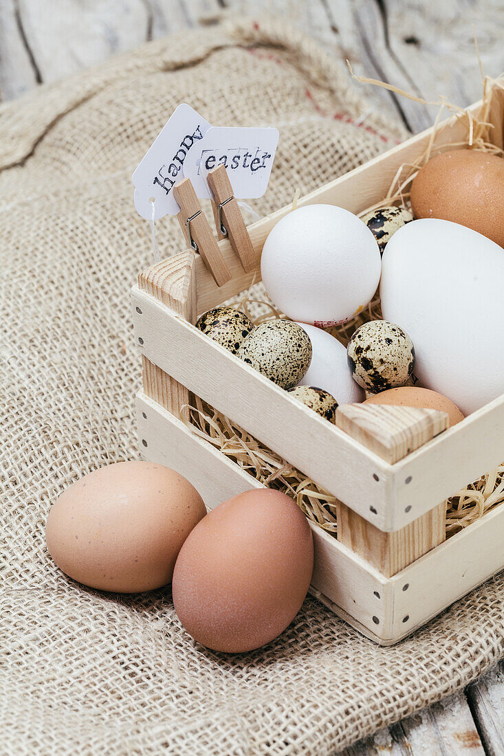 Ein bezauberndes Arrangement von weißen und gesprenkelten Eiern in einem rustikalen Holzkorb mit einem Frohe Ostern"-Anhänger, vor einem Hintergrund aus Sackleinen"
