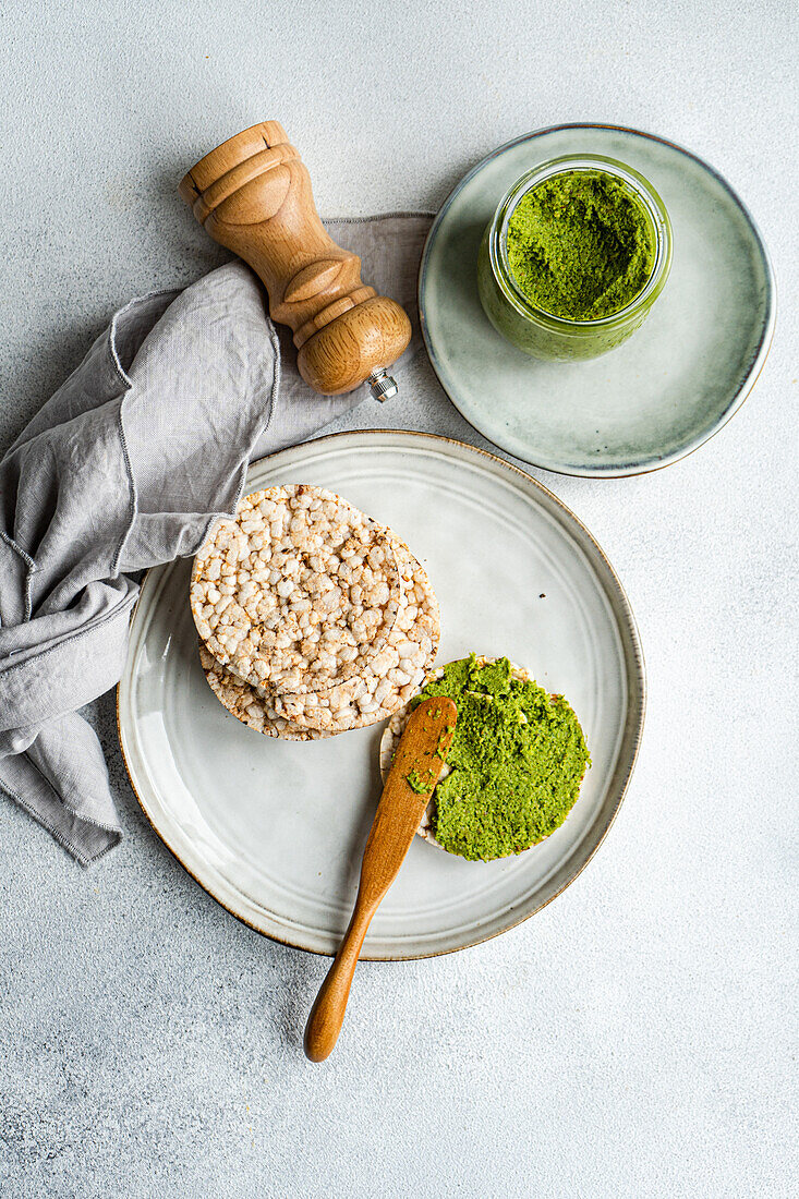 Top view of rice bread on a plate accompanied by a vibrant green spinach pesto pasta-sauce in a glass jar, set against gray backdrop near napkin and salt shaker