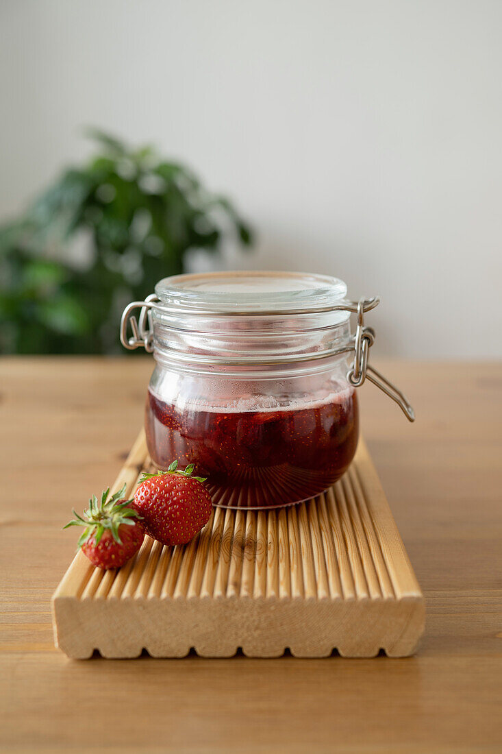 Closeup of glass of strawberry jam with fresh ripe strawberry fruits placed on wooden cutting board over table against blurred background