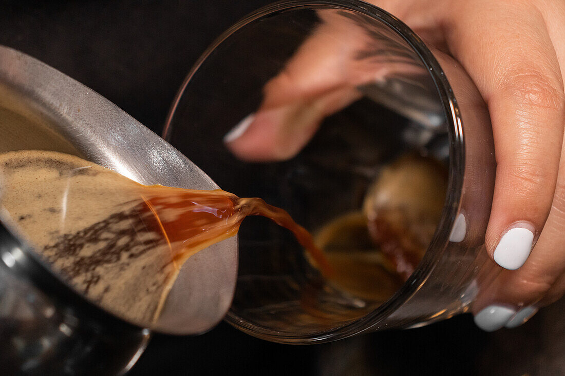 Crop anonymous person pouring coffee from coffeemaker into stylish glass with on counter in cafe